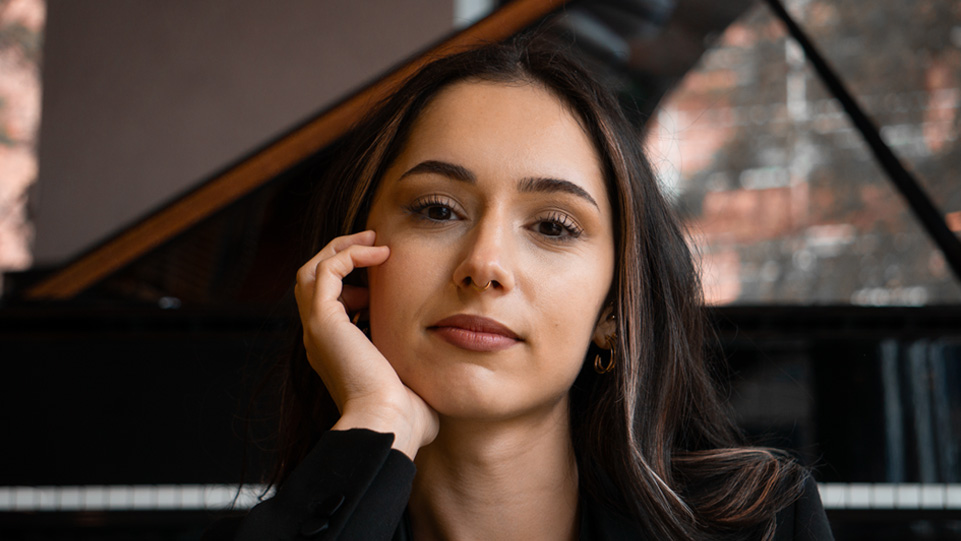 A women with a black tailored top, with a brown and dark brown hair, looking at the camera, with a piano behind her
